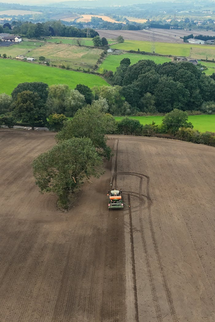 Aerial shot of a tractor plowing a vast field surrounded by lush green landscapes.